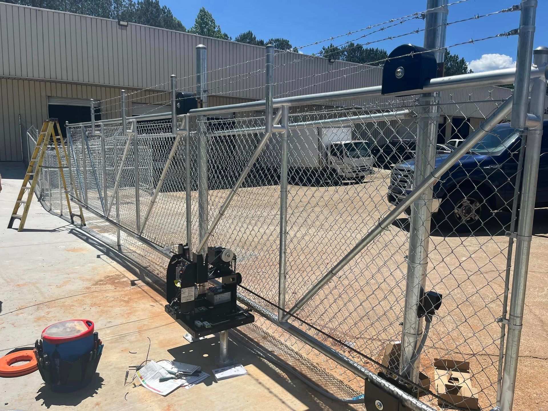 Chain-link fence with barbed wire on top, constructed along building exterior.  Construction equipment nearby, sunny day.