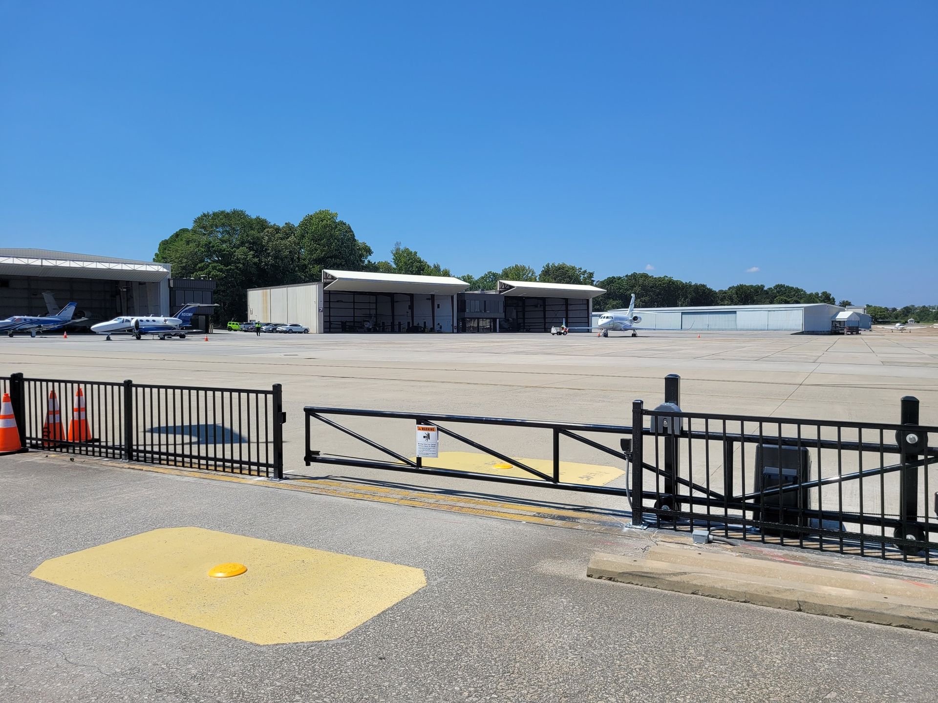 A small airport with parked airplanes, hangars, and a gated entrance under a clear blue sky.