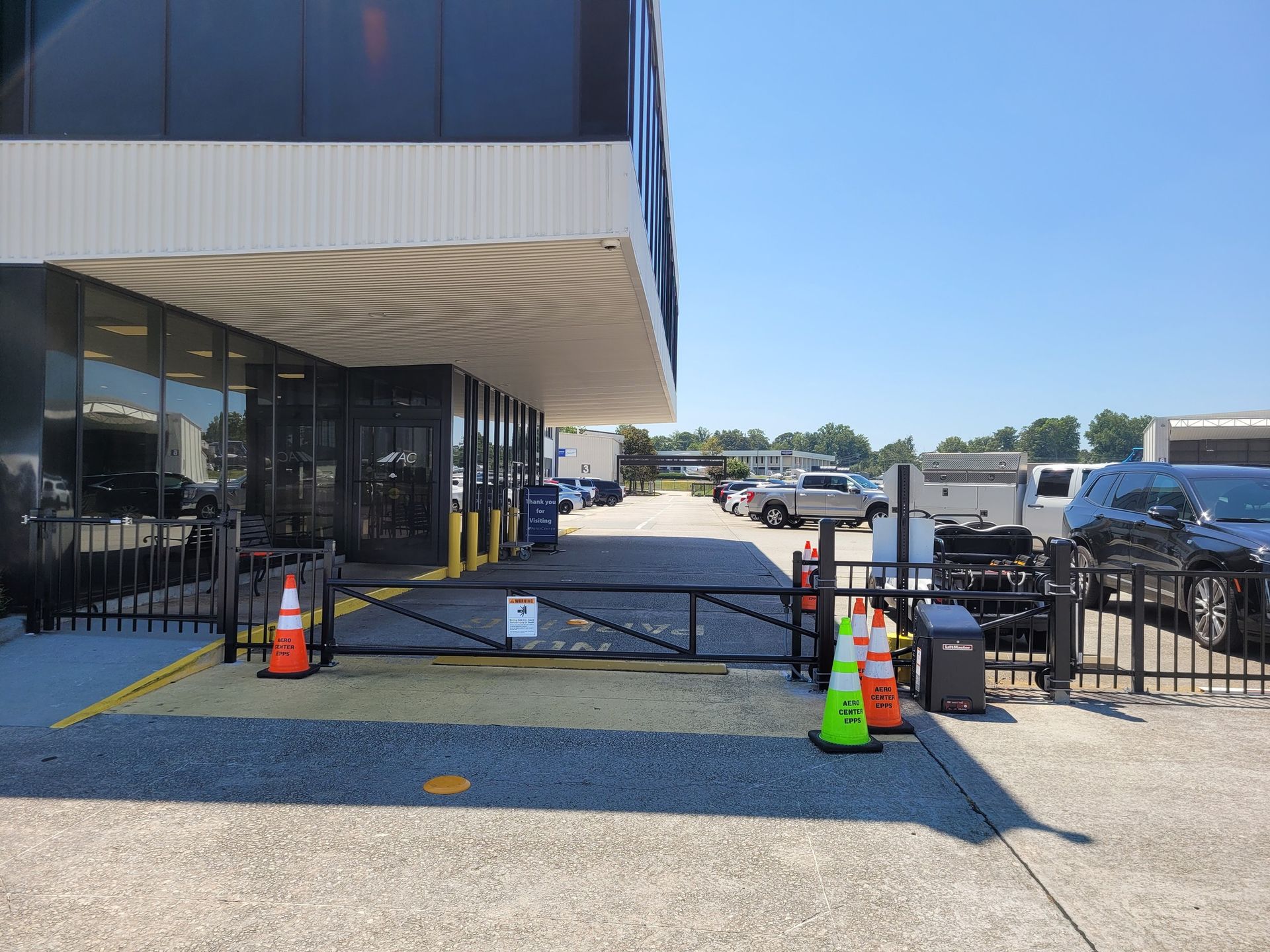 Black gate at building entrance, orange traffic cones, vehicles parked beyond, clear sky.