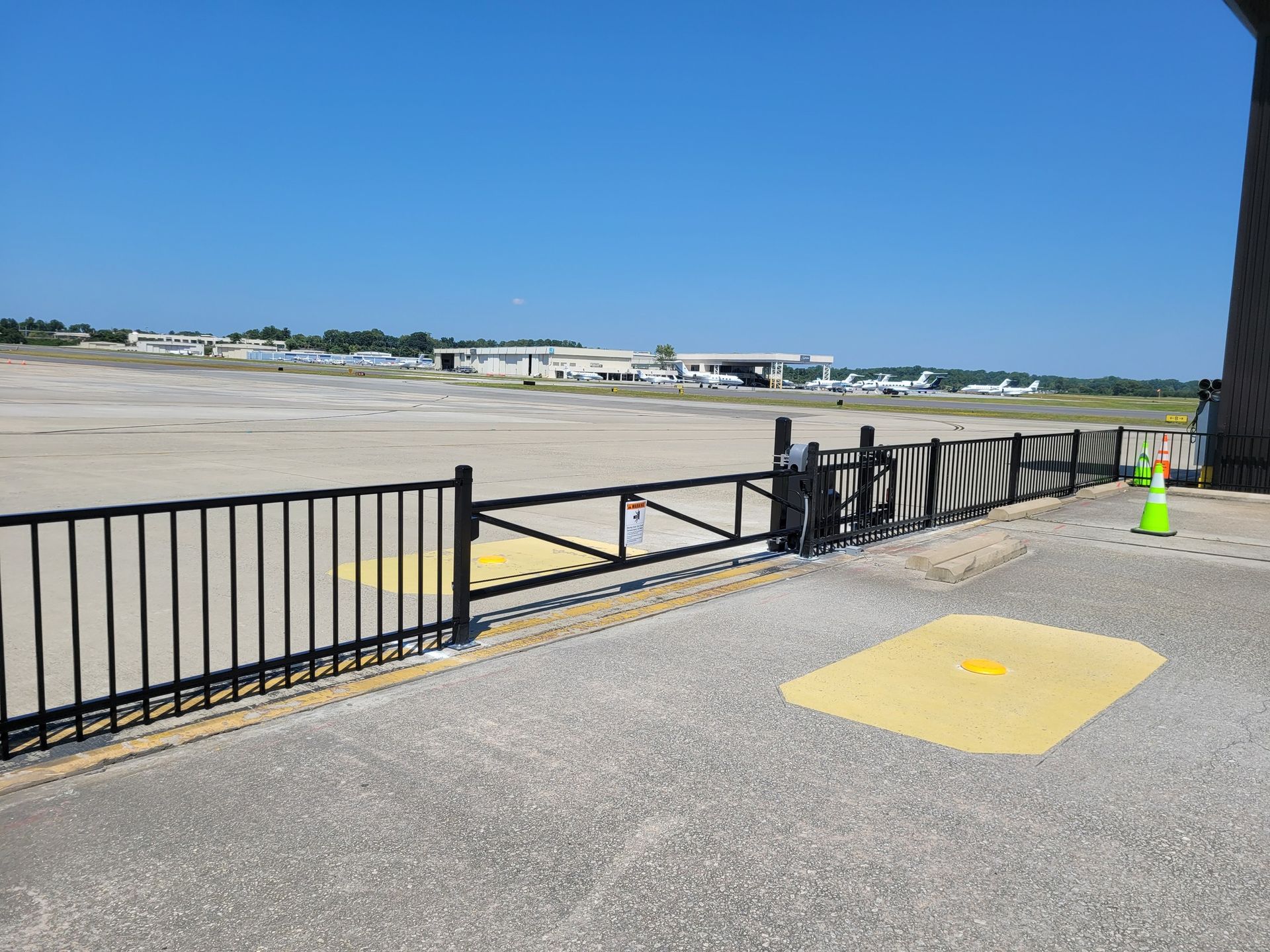 View of an airport tarmac from a paved walkway, black fence, blue sky.