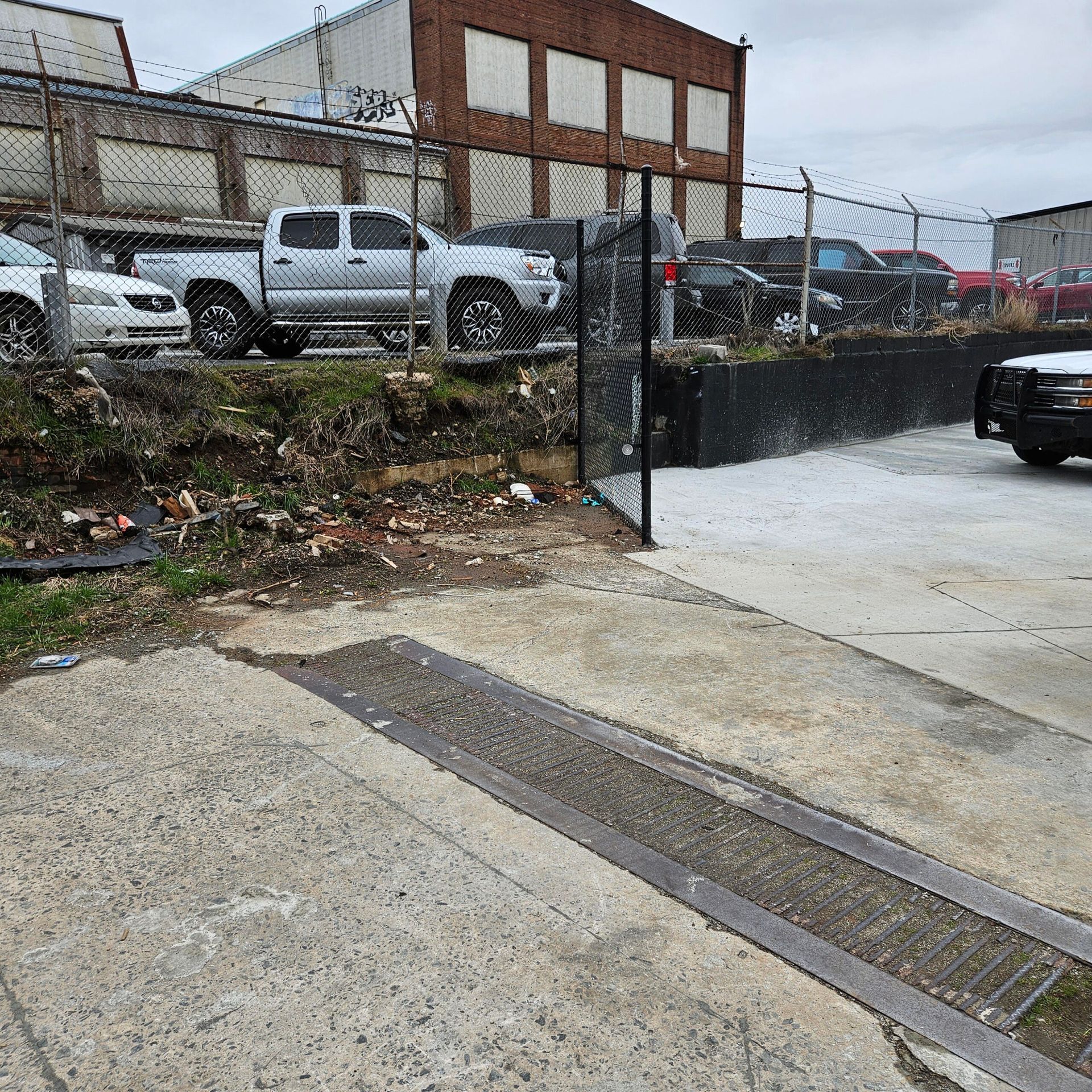 Concrete area next to a fenced lot with cars. A pickup truck, building in the background. Cloudy day.