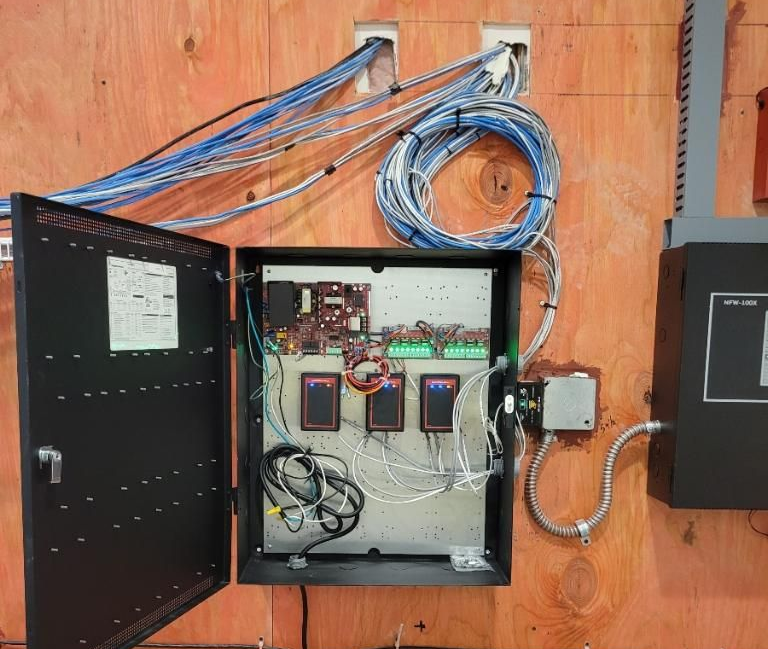 Open black electrical box with circuit boards, wiring, and three card readers mounted on wood paneling.