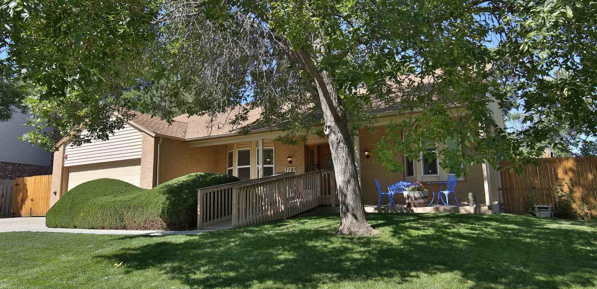 A one-story house with a wooden ramp and a tree in the front yard on a sunny day.