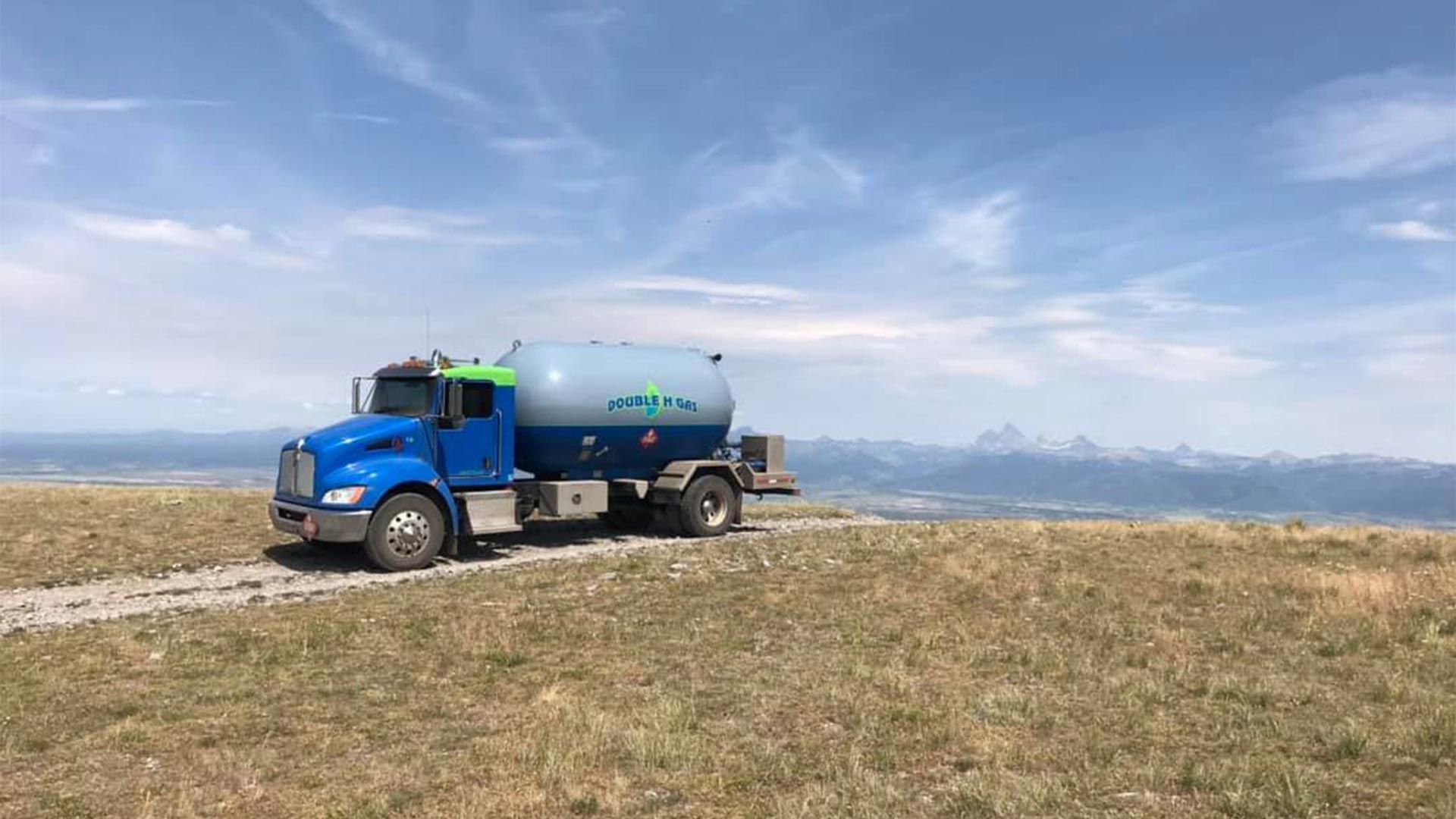 A blue truck is driving down a dirt road on top of a grassy hill.