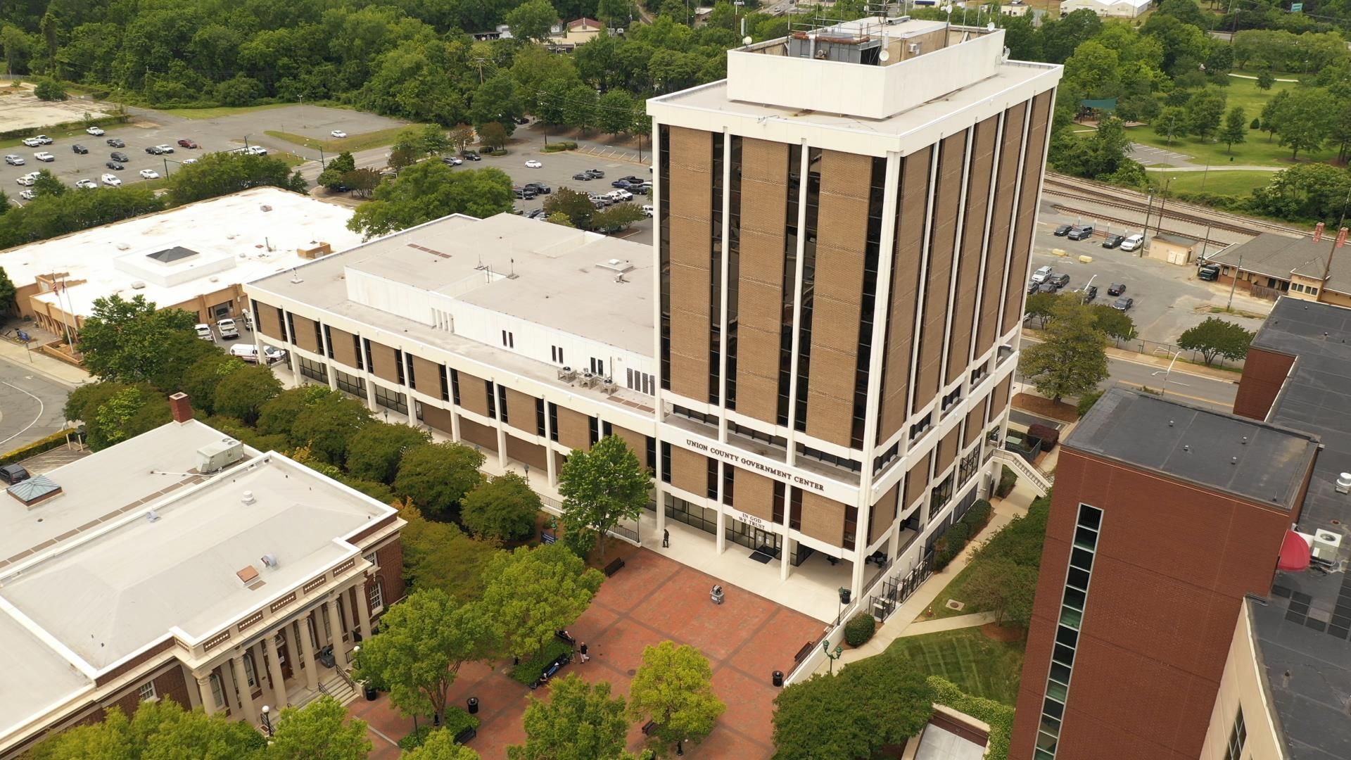 Aerial view of a tall, brown and white office building with adjacent low-rise structures and a plaza with trees.