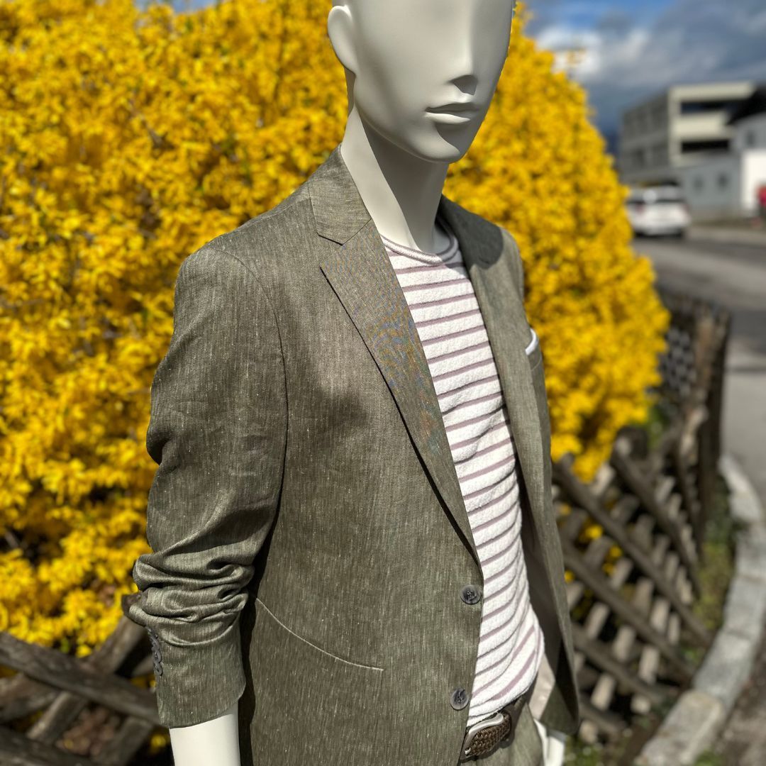 Eine Schaufensterpuppe mit Anzugjacke und gestreiftem Hemd steht vor einem gelben Busch.