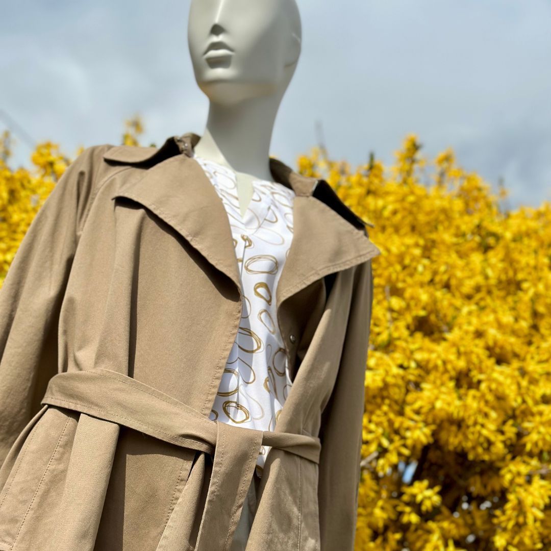 Eine Schaufensterpuppe im Trenchcoat steht vor gelben Blumen