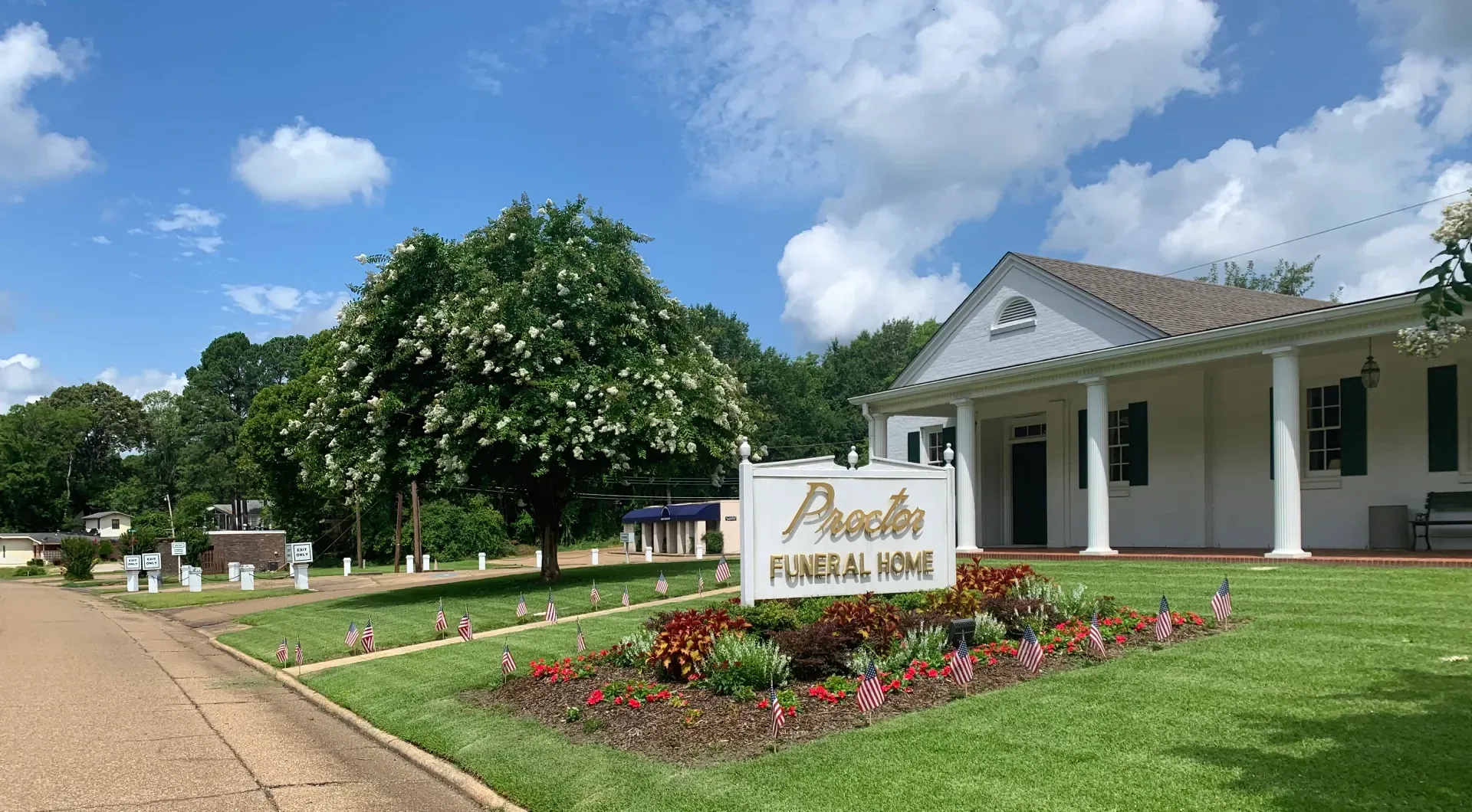 White clubhouse entrance with sign, flower bed, green lawn, and trees under a blue sky