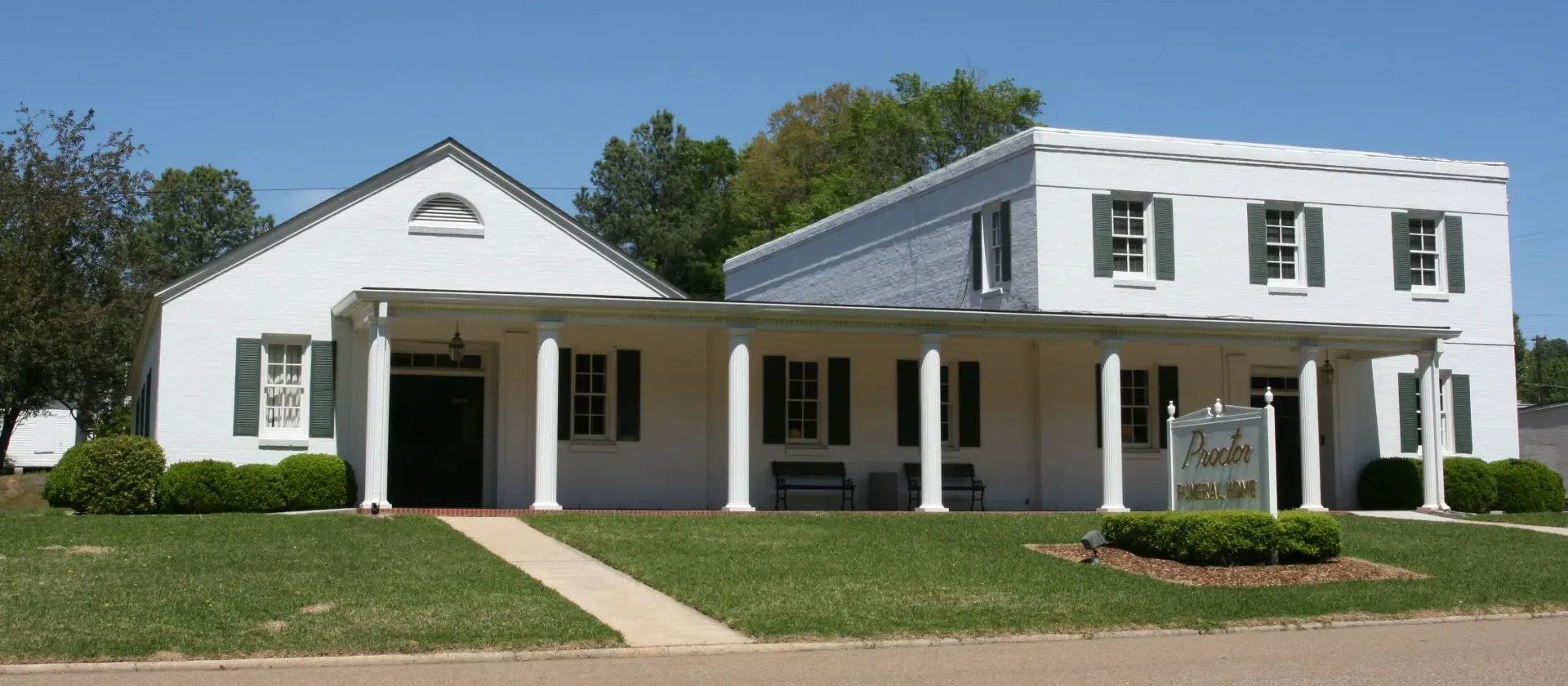 White two-story building with porch and columns, set behind a lawn and walkway on a sunny day