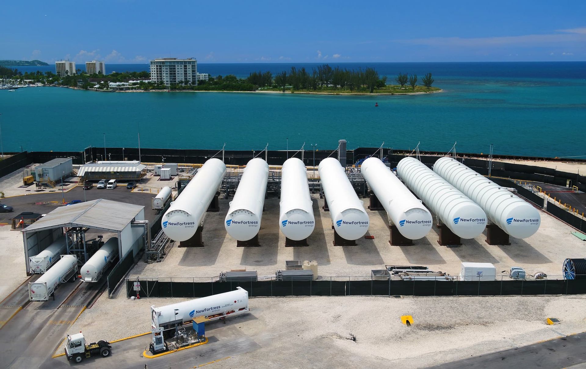 Coastal industrial site with rows of white storage tanks beside turquoise water and sandy shore