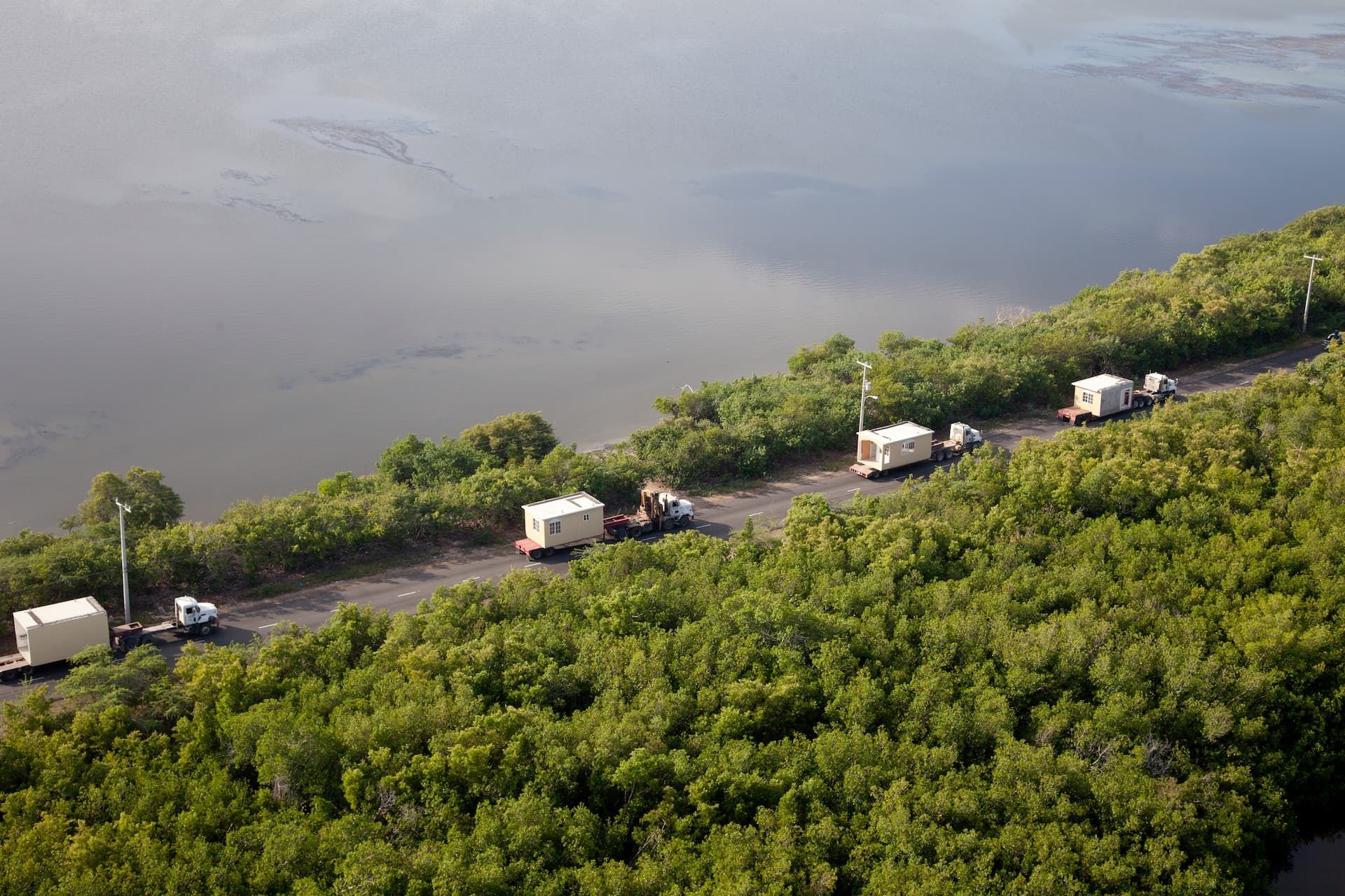 Line of white trucks driving along a narrow road beside water and dense green hillside.