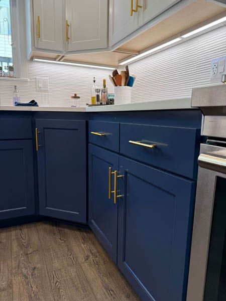 Modern kitchen corner with navy blue cabinets, gold handles, white backsplash, and wood-look floor