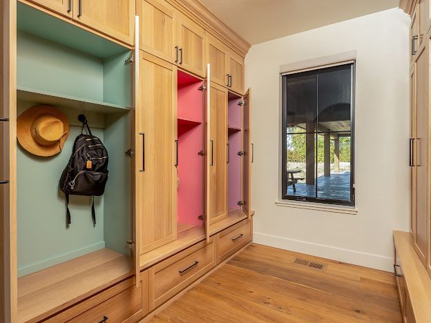 Built-in wooden closet with open pastel cubbies, hat and backpack, beside a window in a bright room