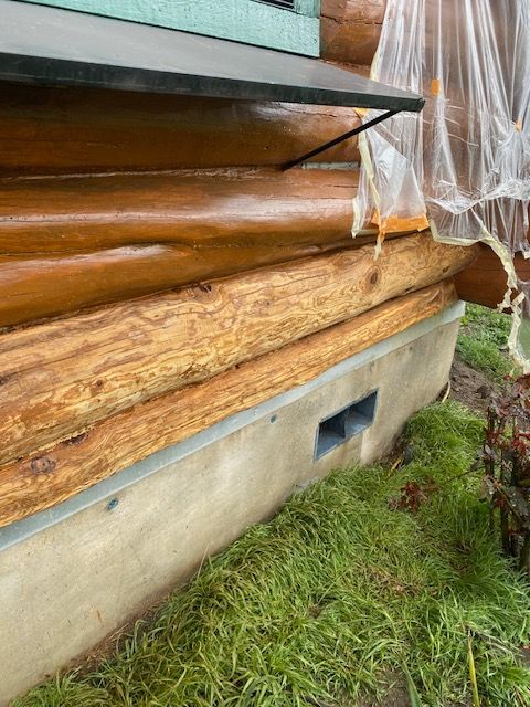 Log cabin wall with exposed insulation, plastic sheeting, and a concrete foundation above grass