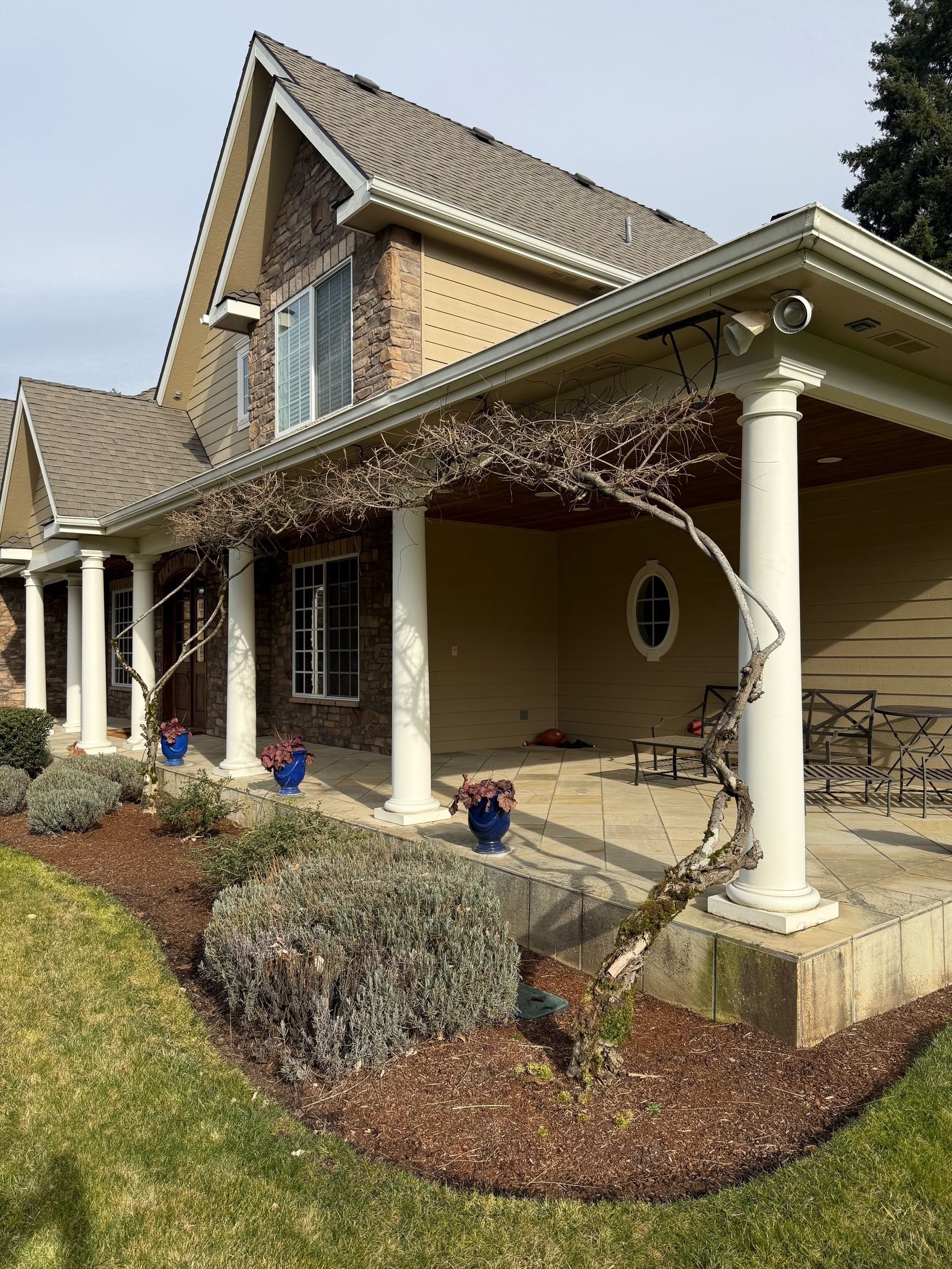 Front porch of a beige brick house with white columns, gabled roof, and winter landscaping.