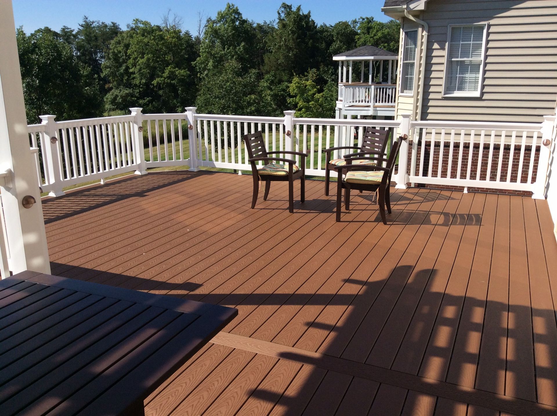 Wooden deck with chairs and table, house in the background, surrounded by trees under a blue sky.