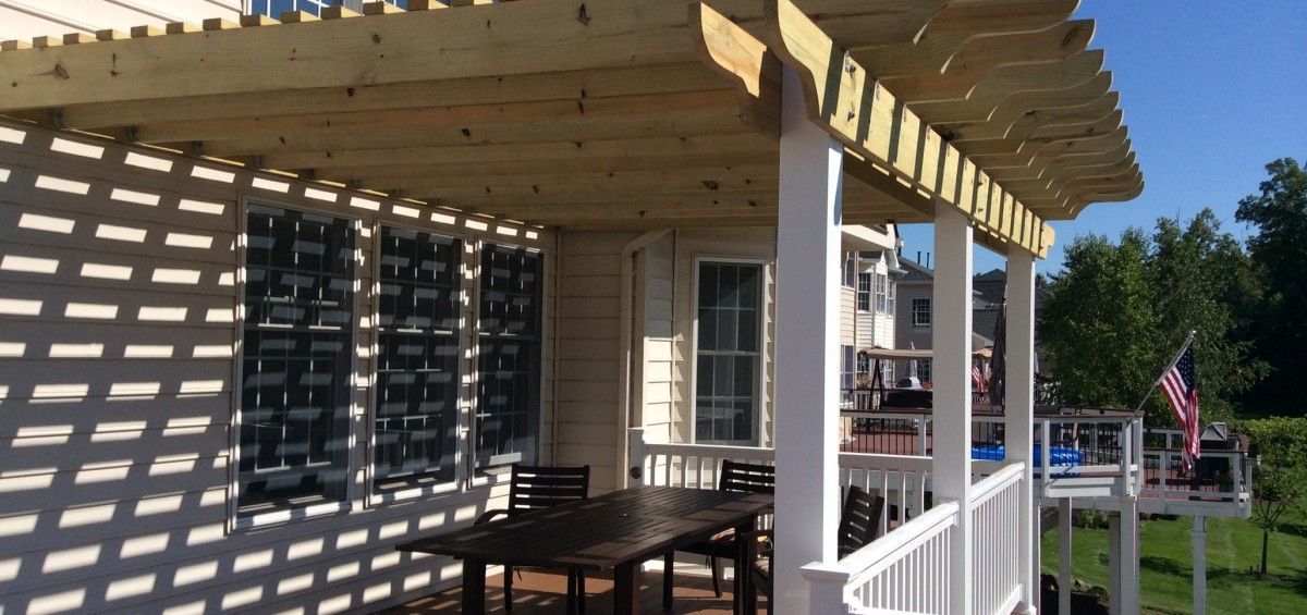 Wooden pergola over a porch with a table, chairs, and white railing. American flag in the background.