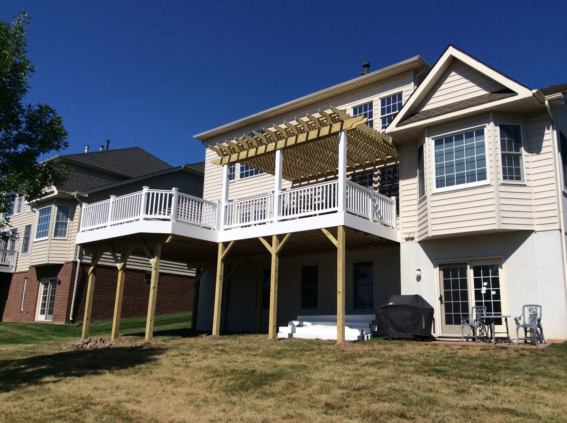 Two-story house with a wooden deck and pergola in a grassy backyard under a clear blue sky.