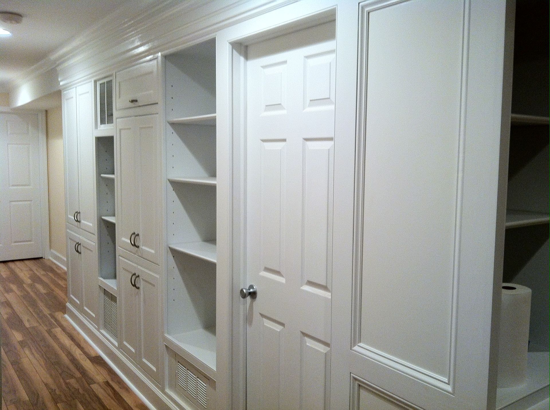 Built-in white cabinets with shelves, a door, and trim along a hallway with wood flooring.