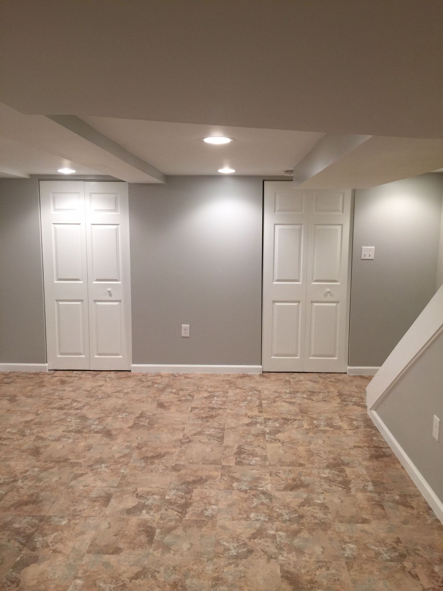 A finished basement with grey walls, two white bi-fold doors, recessed lighting, and patterned flooring.