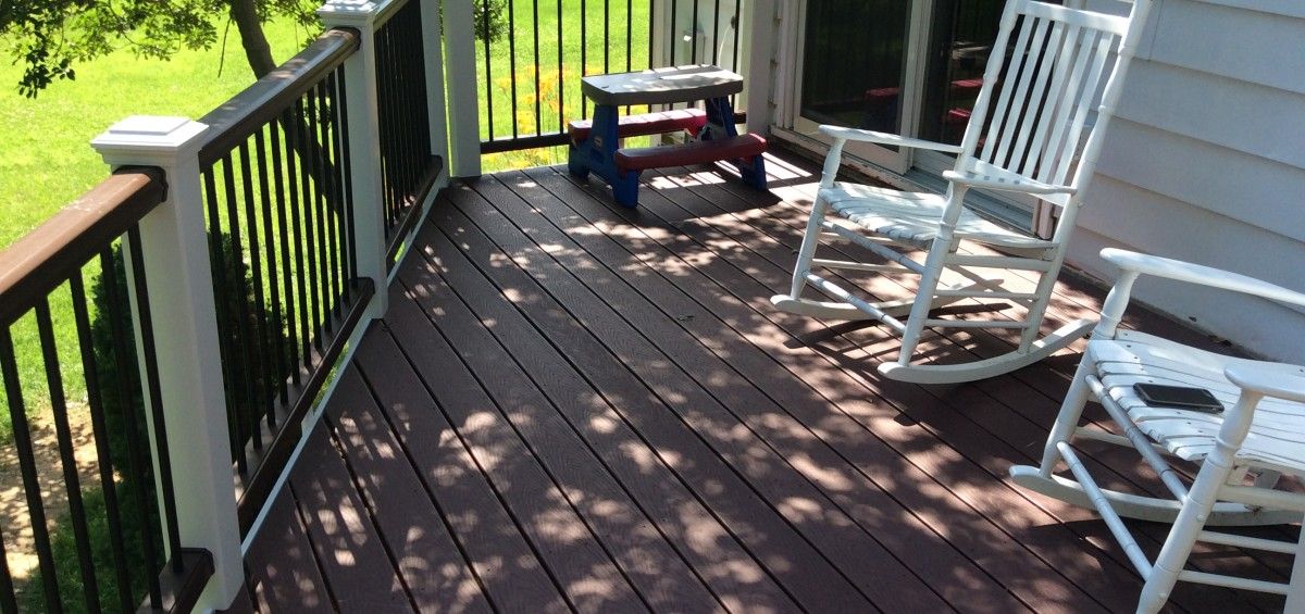 A wooden deck with white rocking chairs and a small bench. Deck railing is black and brown. Sunny day.
