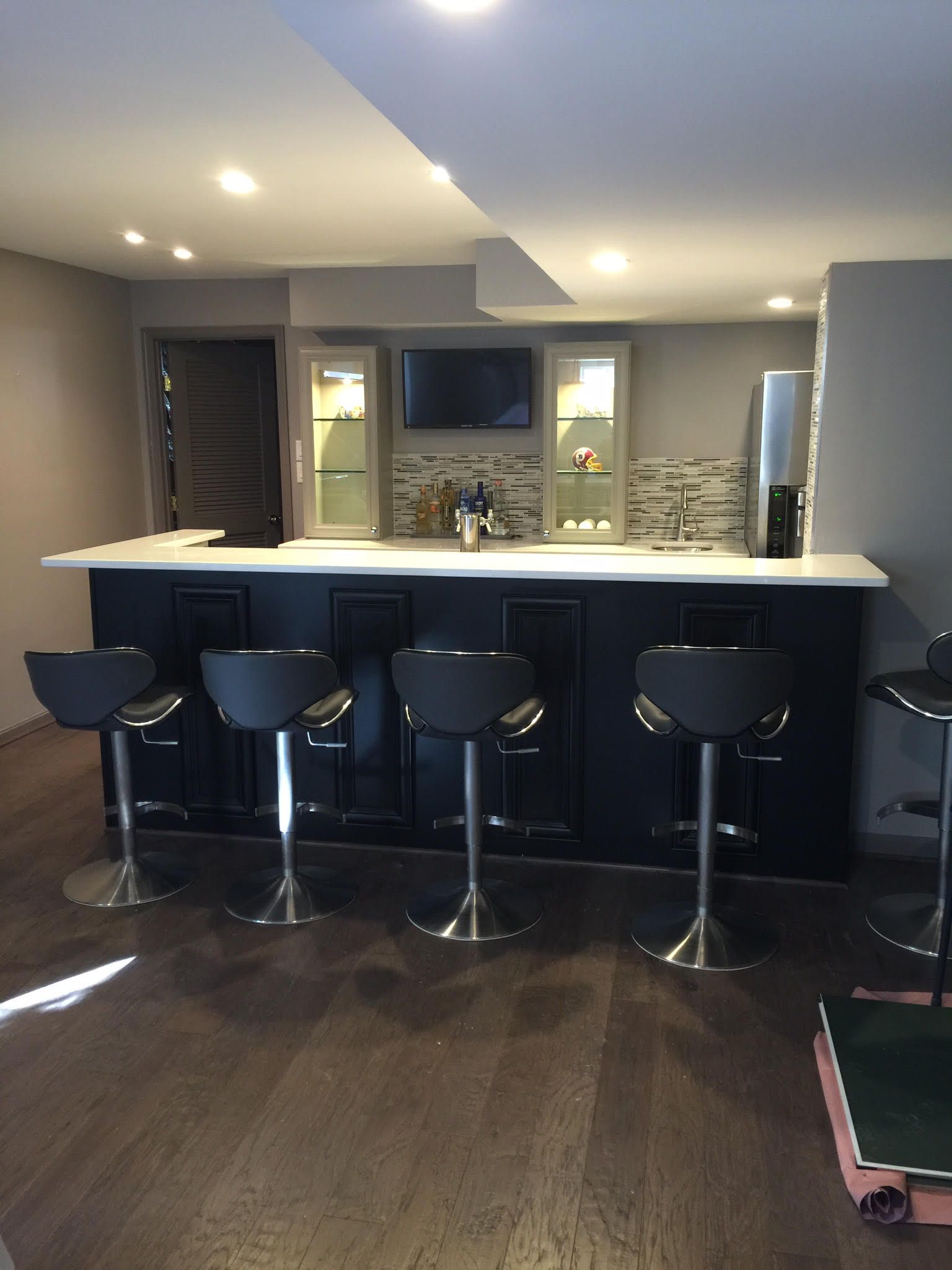 Basement bar with black cabinets, white countertop, stools, TV, and cabinets with illuminated shelves.