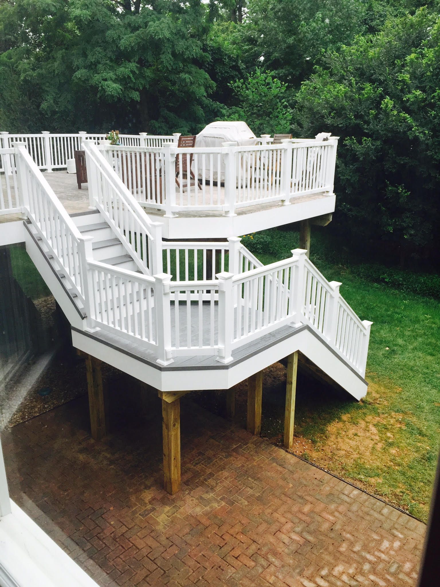 Multi-level white deck with stairs, surrounded by greenery. Brick patio below.