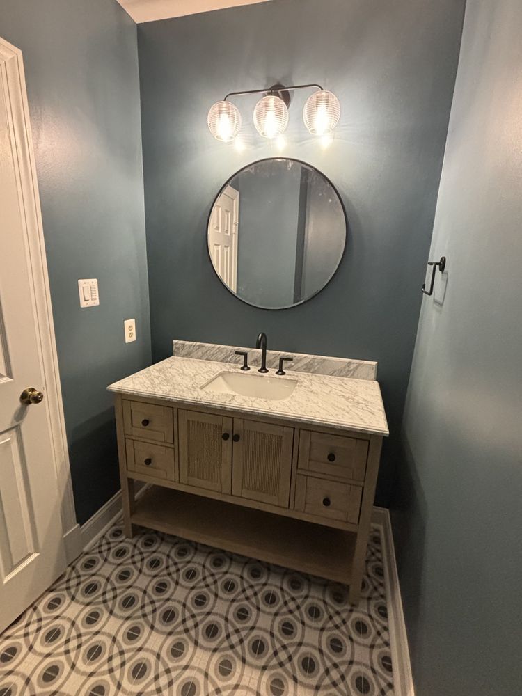 Bathroom with vanity, round mirror, and patterned floor. Light blue walls, wooden vanity, marble countertop.