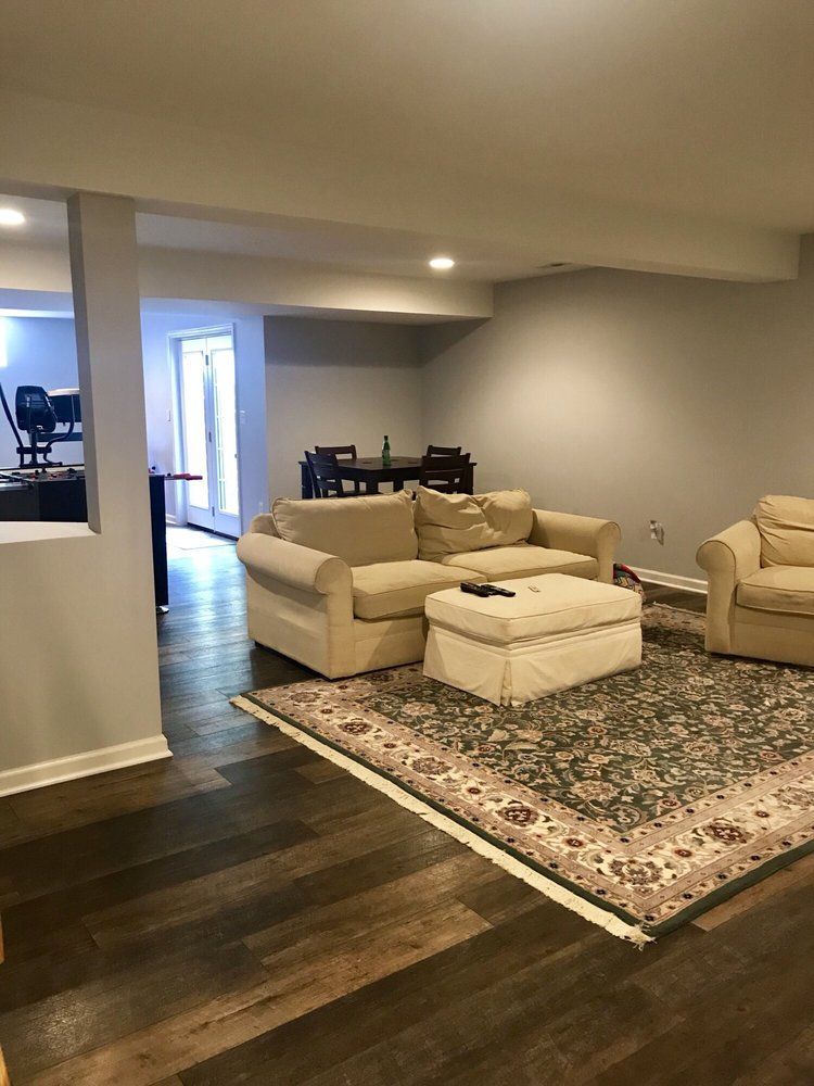 Cozy living room with a beige sofa, ottoman, and patterned rug. Dark wood floors, a dining area in the background.