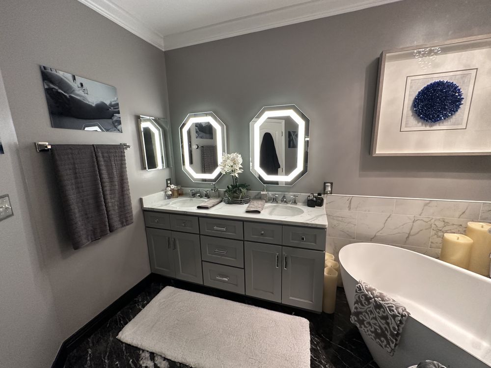 Gray bathroom with double sinks, lighted mirrors, tub, and artwork.