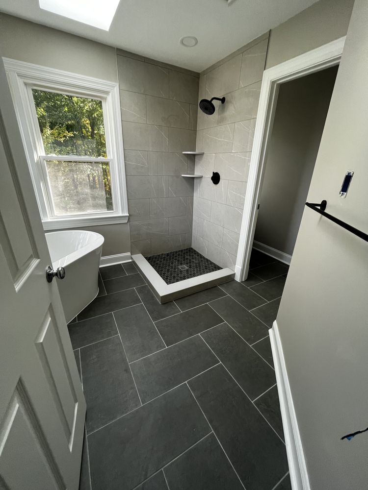 Modern bathroom with gray tile, walk-in shower, freestanding tub near window, and dark tile floor.