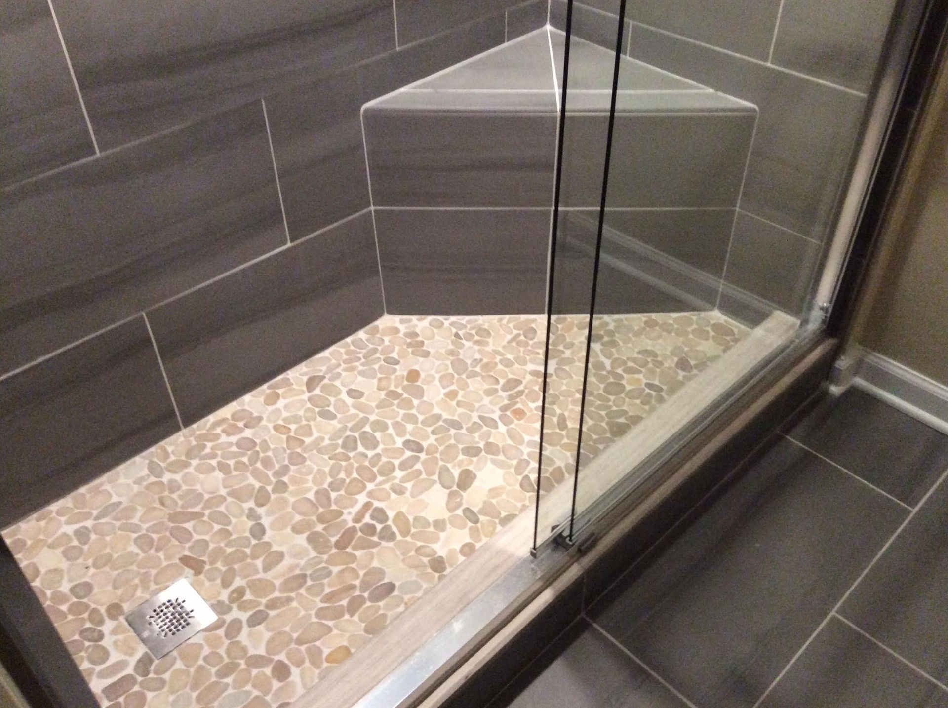 Shower with pebble floor, gray tile walls, and triangular shelf. Glass door and chrome drain visible.