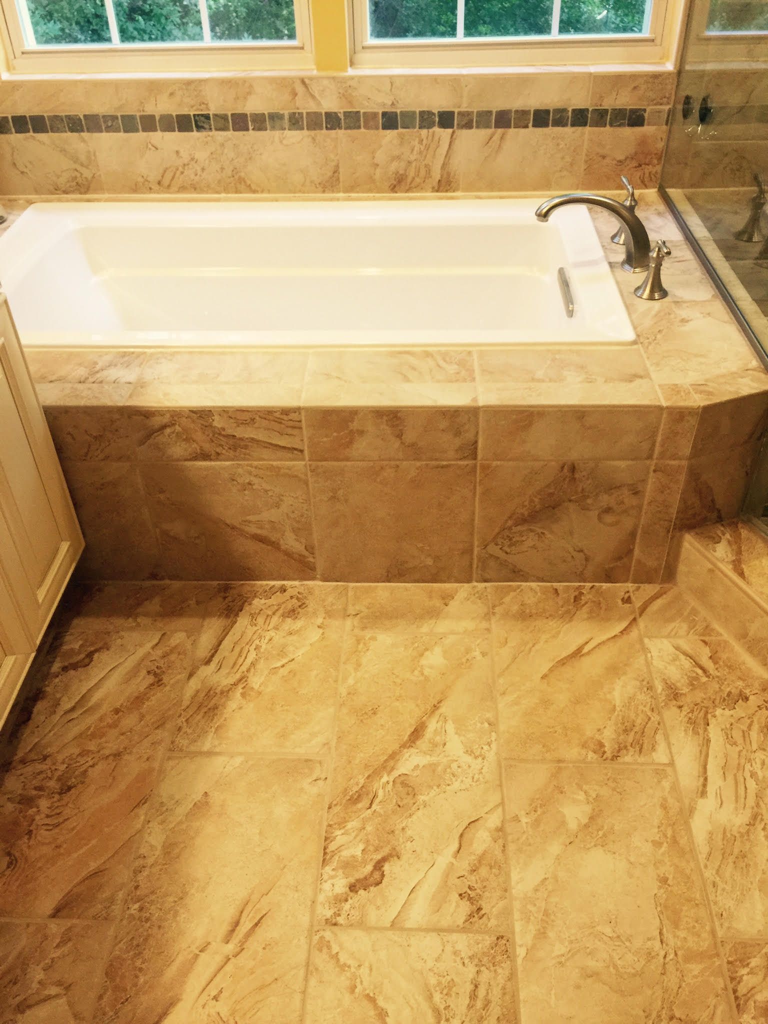 Bathroom with a white bathtub surrounded by tan tiles and a window in the background.