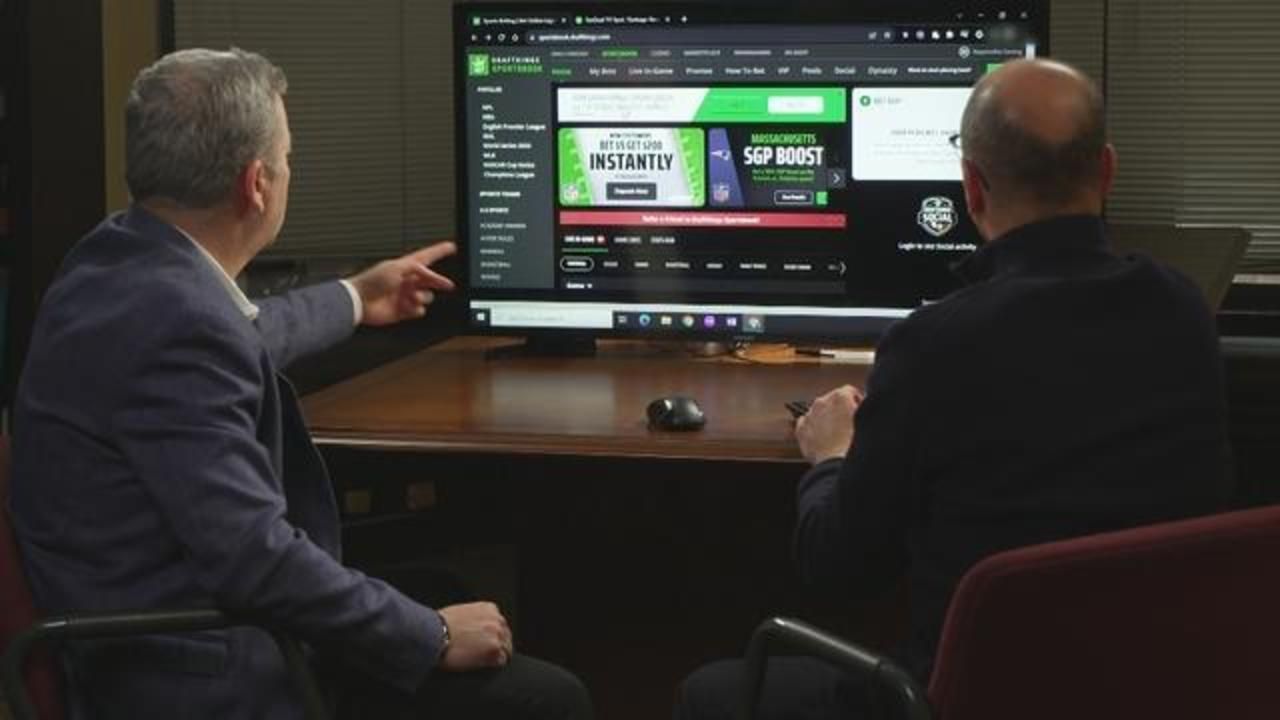 Two people in a dim office point at a large monitor showing a betting or casino website.