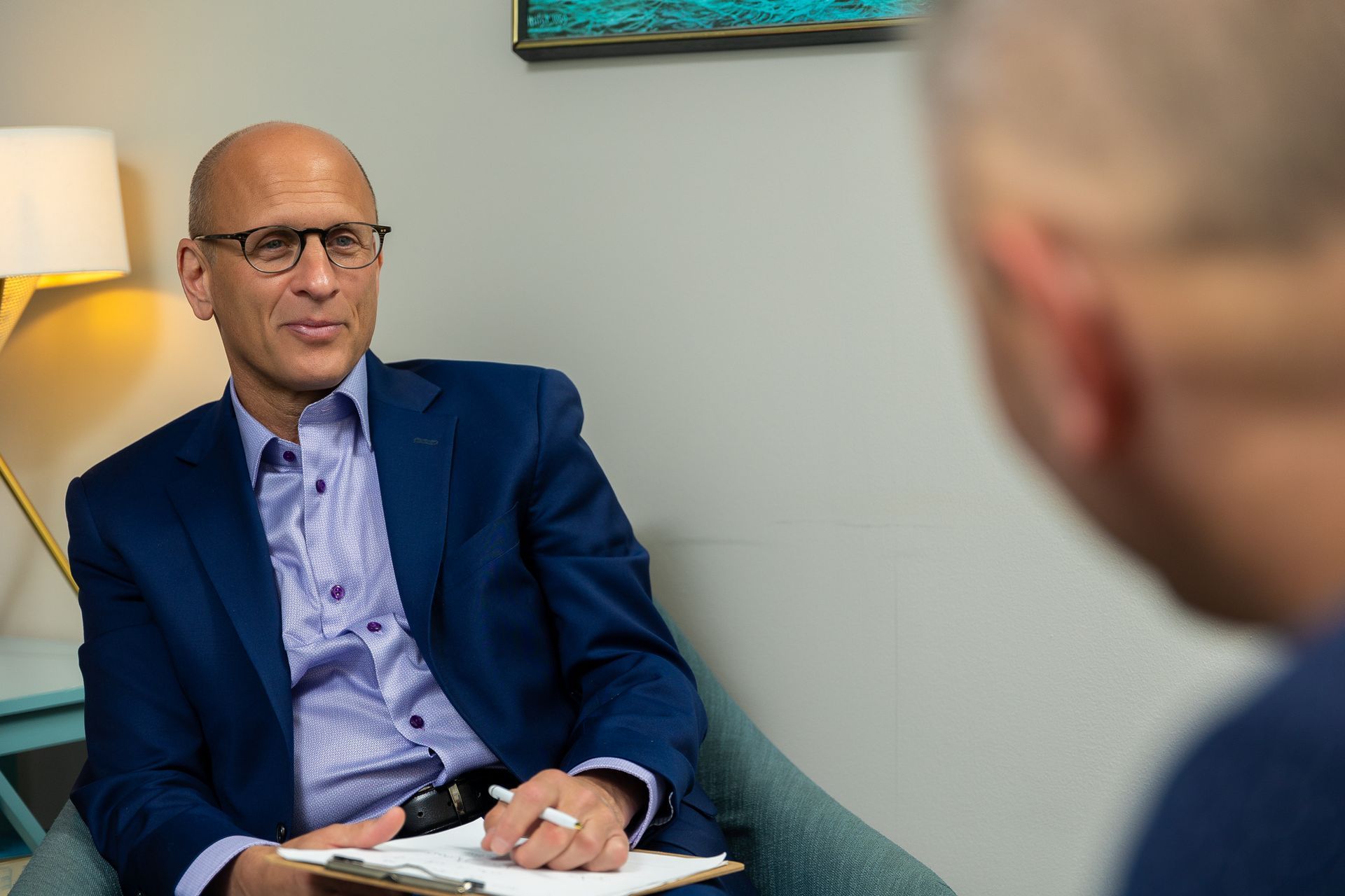 Man in blue blazer speaking in a meeting room, with another person blurred in the foreground.