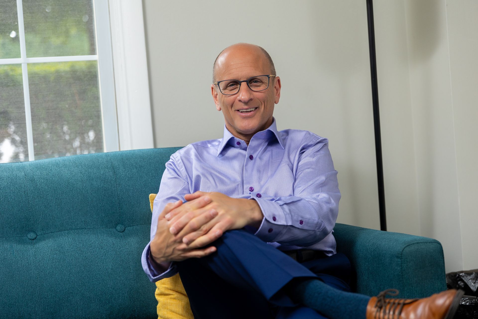 Smiling man in a blue shirt sitting on a teal couch with hands clasped, in a bright room.