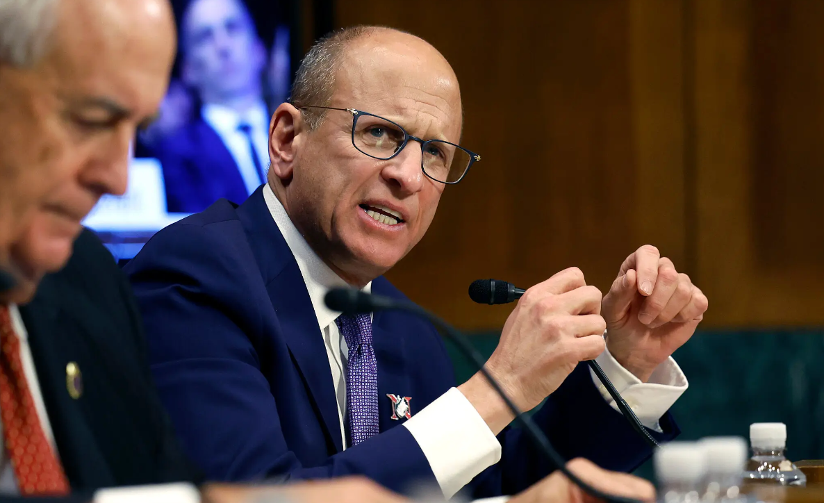 Man speaking emphatically at a hearing table, gesturing with his hands and wearing glasses and a suit.