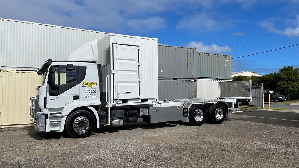 A White Semi Truck is Parked in Front of a Building Filled With Shipping Containers — Davis Removals & Storage in Chermside, QLD