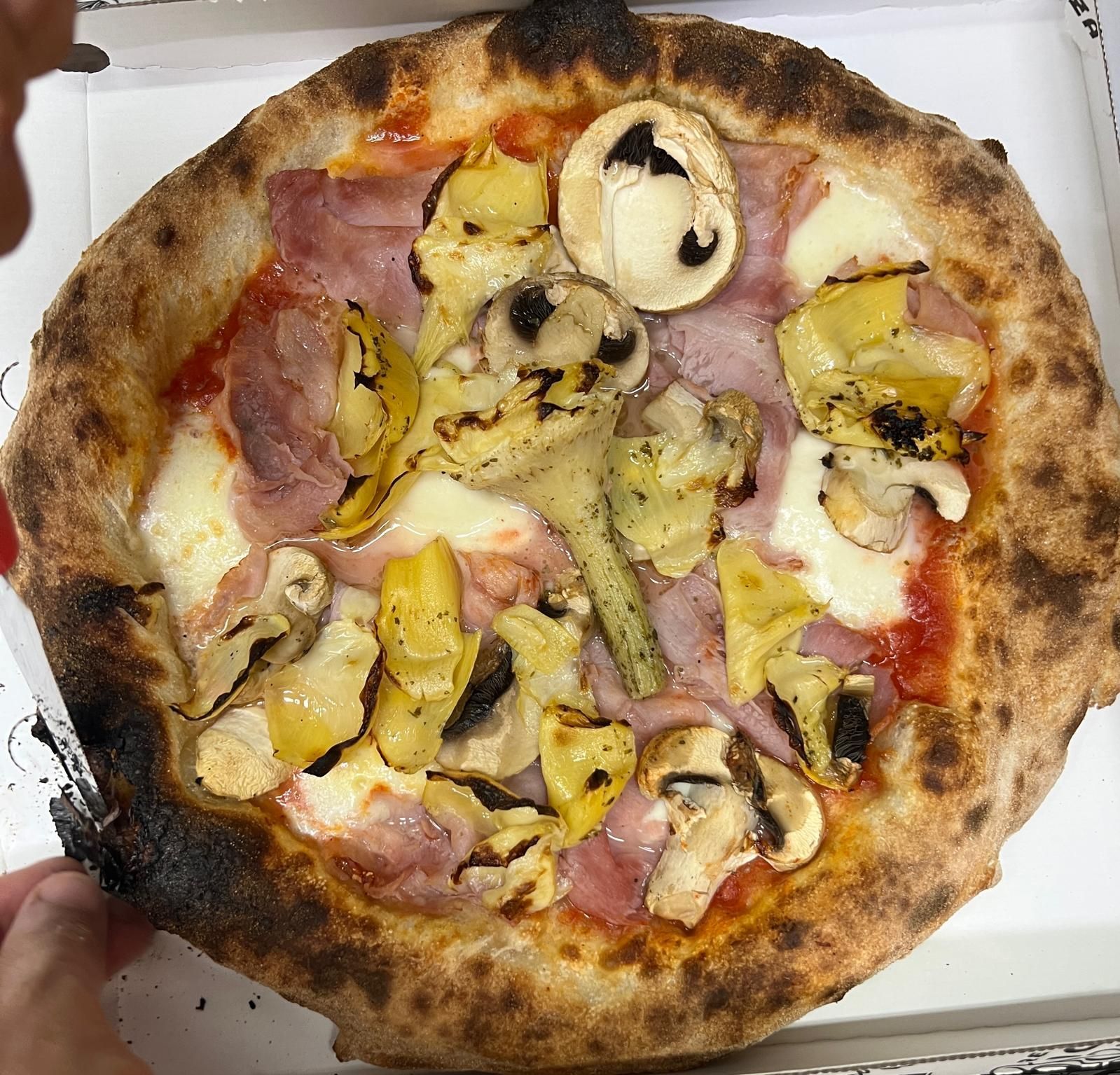 Scatola della pizza aperta con una pizza dalla crosta bruciacchiata condita con prosciutto, funghi, carciofi e formaggio.