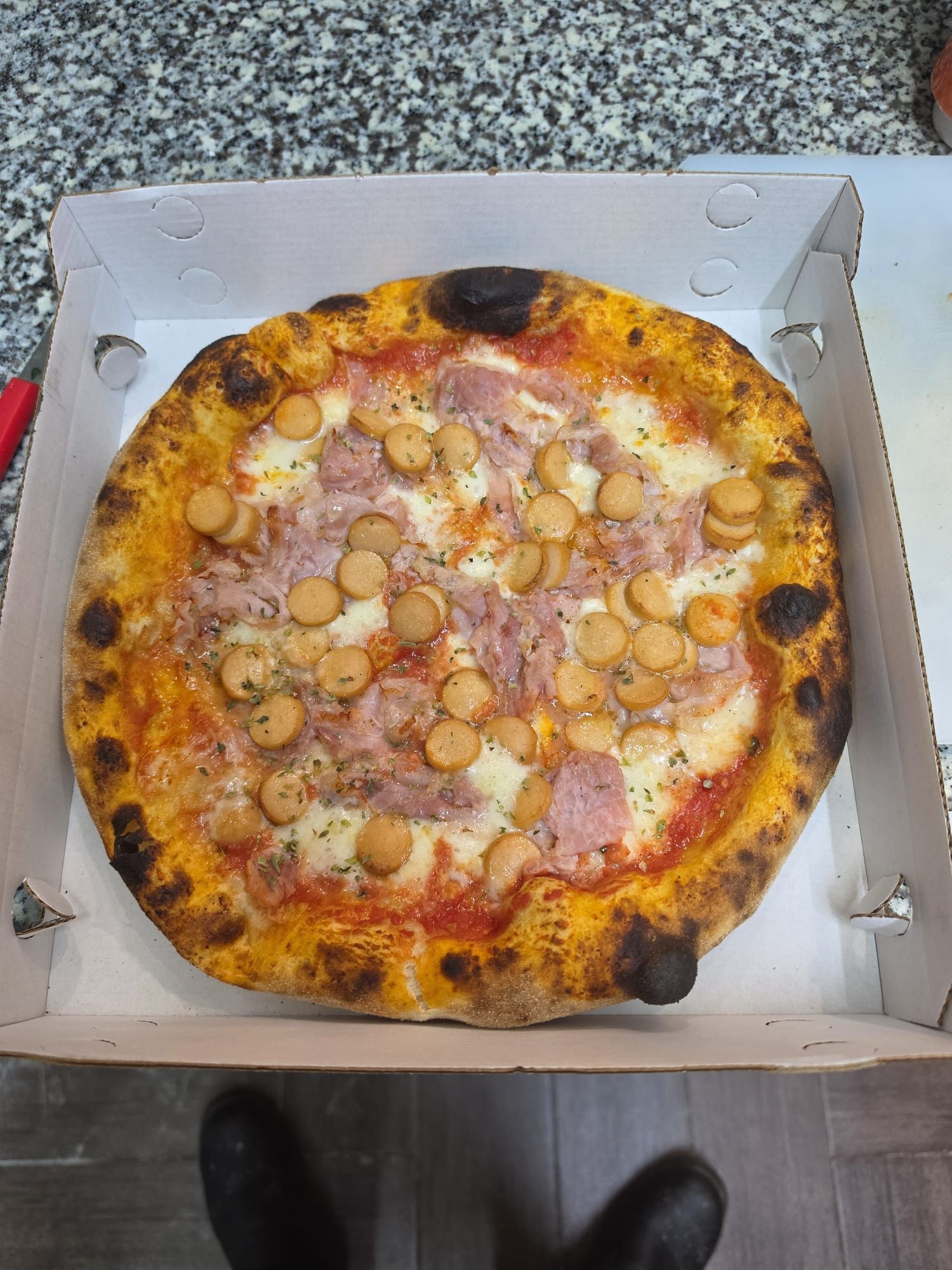 Pizza aperta in una scatola di cartone, condita con frutti di mare e formaggio, su un pavimento grigio.