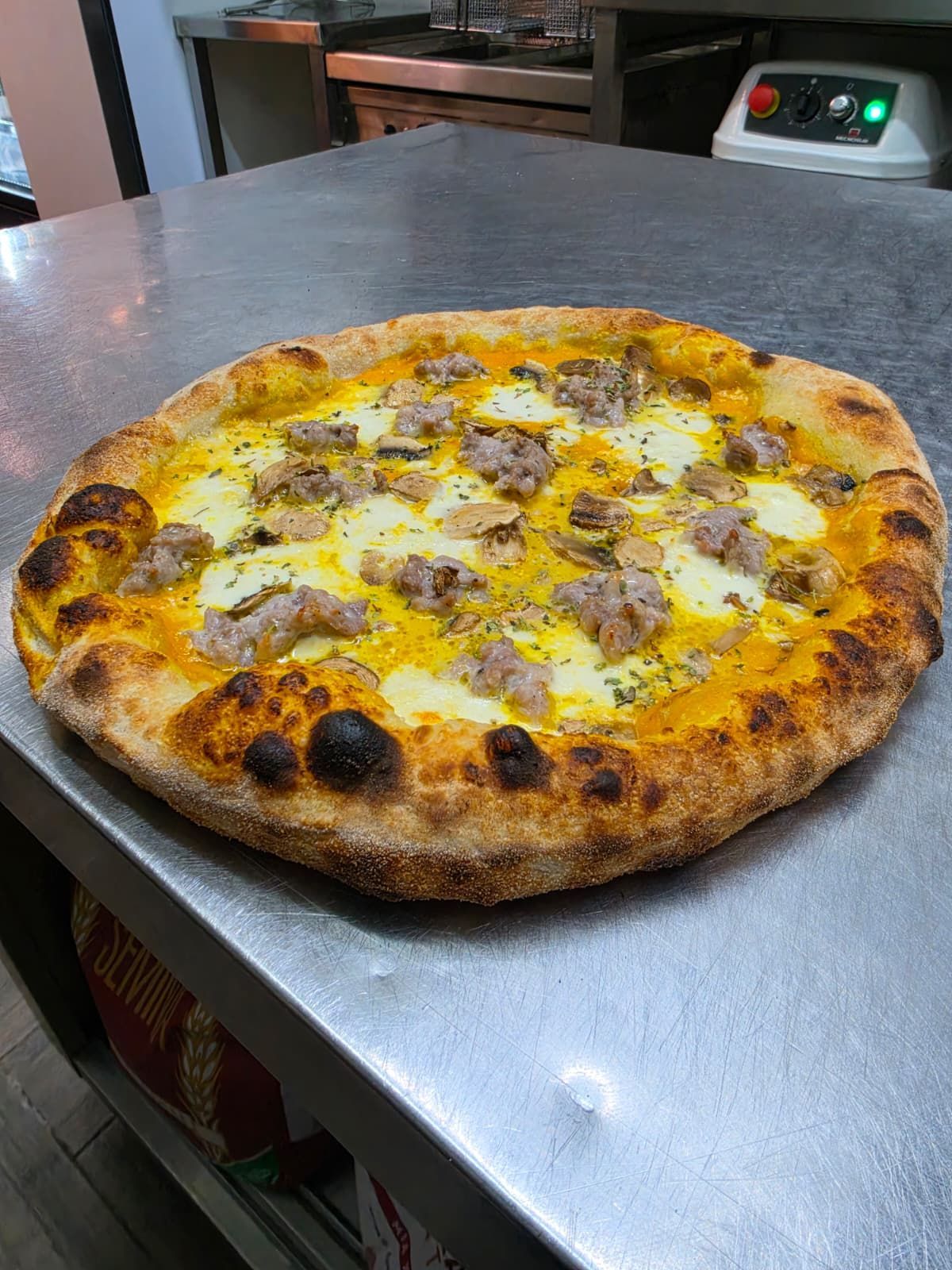 Pizza al formaggio su un tavolo di metallo nella cucina di un ristorante, con una crosta dorata e punti bruciacchiati.