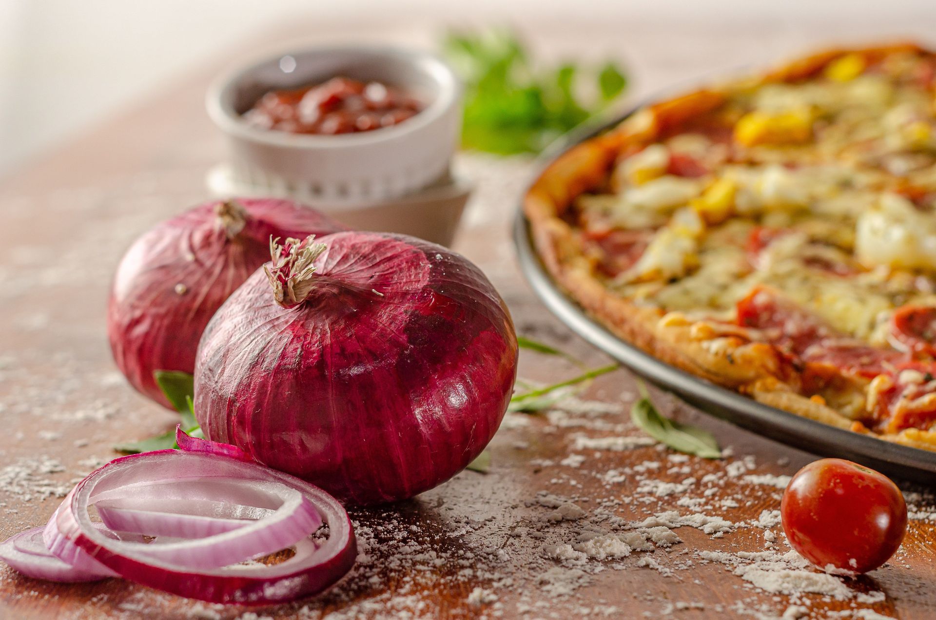 Cipolle rosse su un tavolo rustico accanto a una pizza con pomodori e una ciotola di spezie.