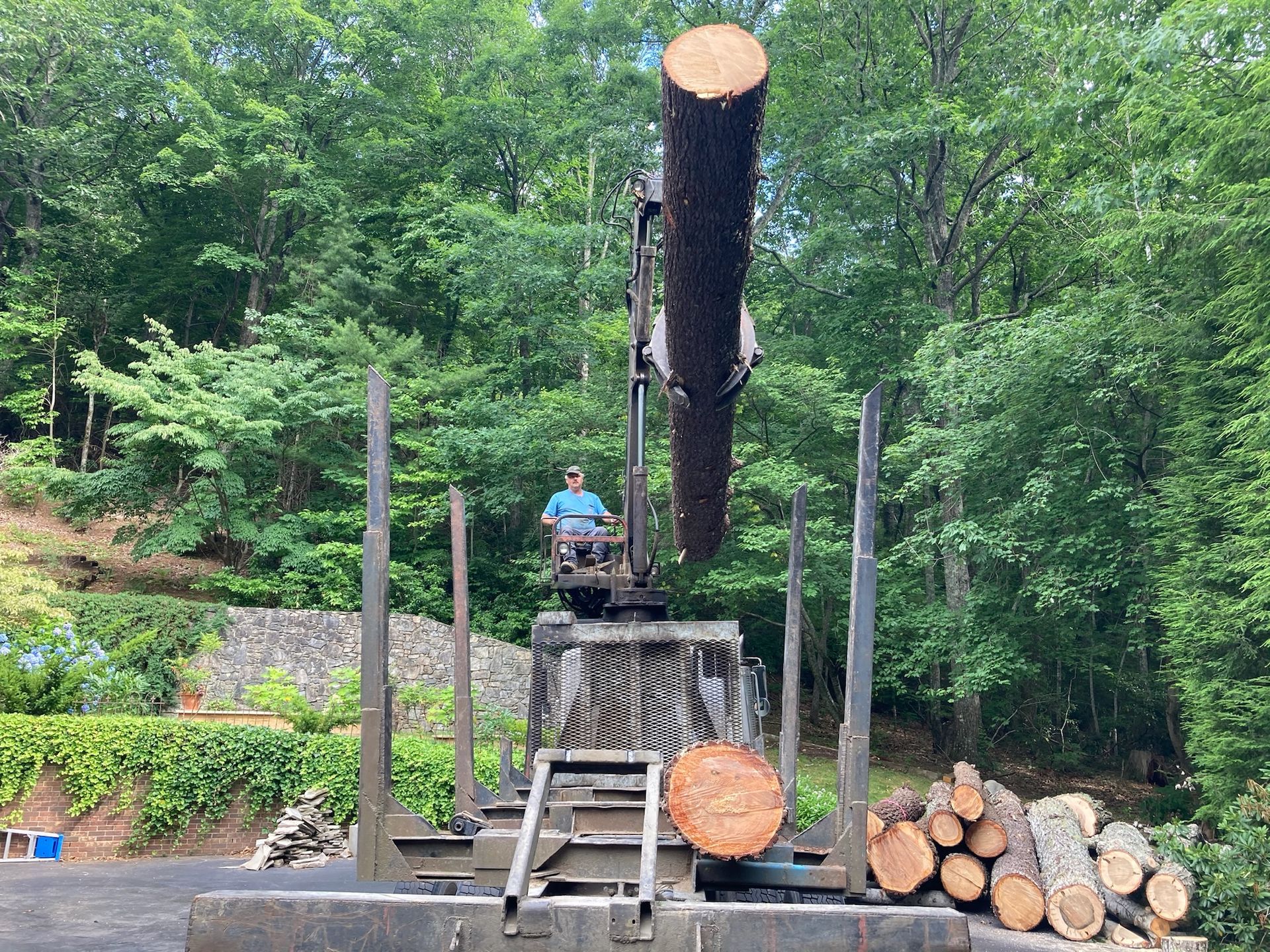 Man operating a crane lifting a log into a truck bed, surrounded by trees and firewood.