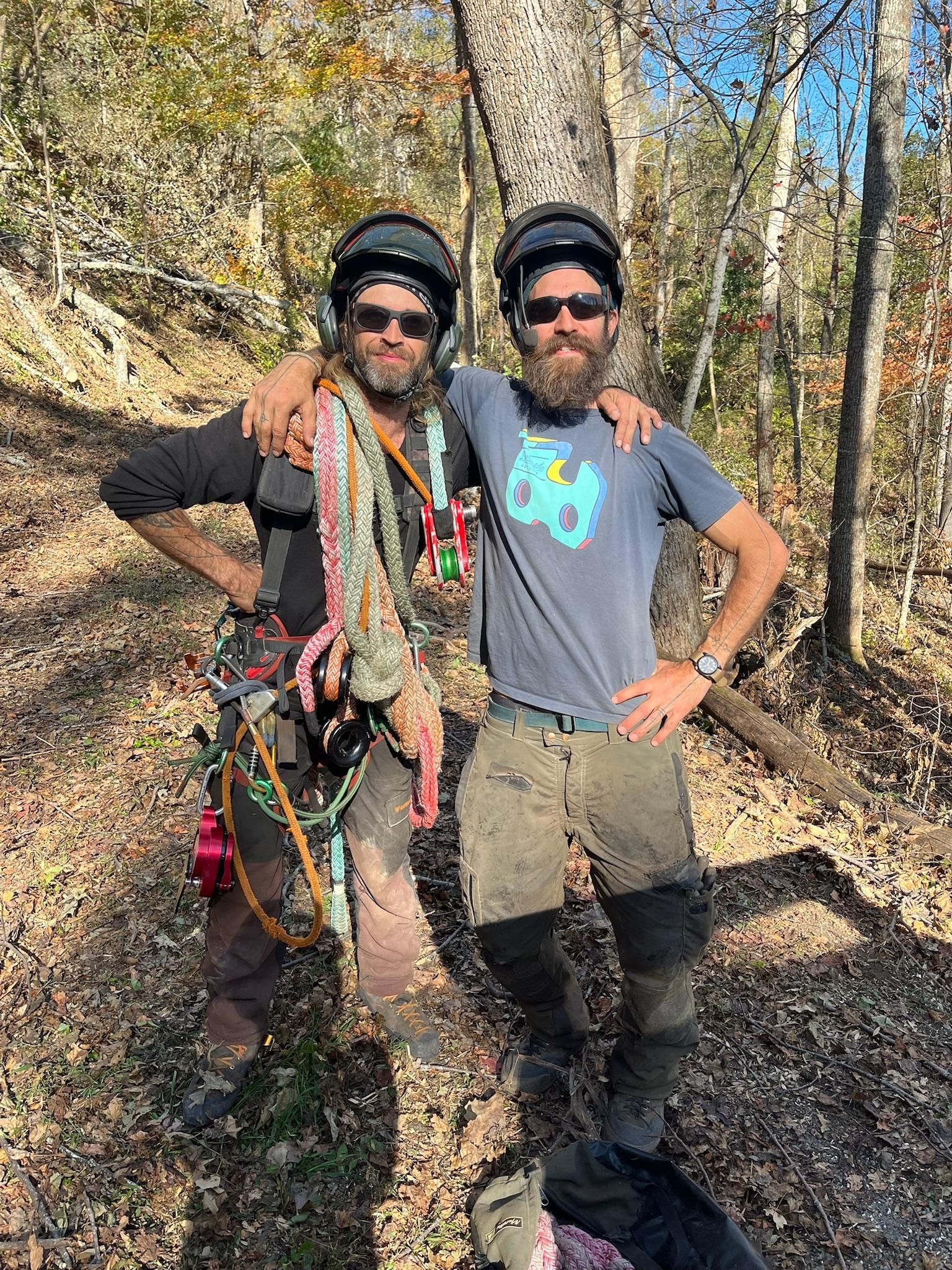 Two men in helmets and harnesses pose in a wooded area, arms around each other.