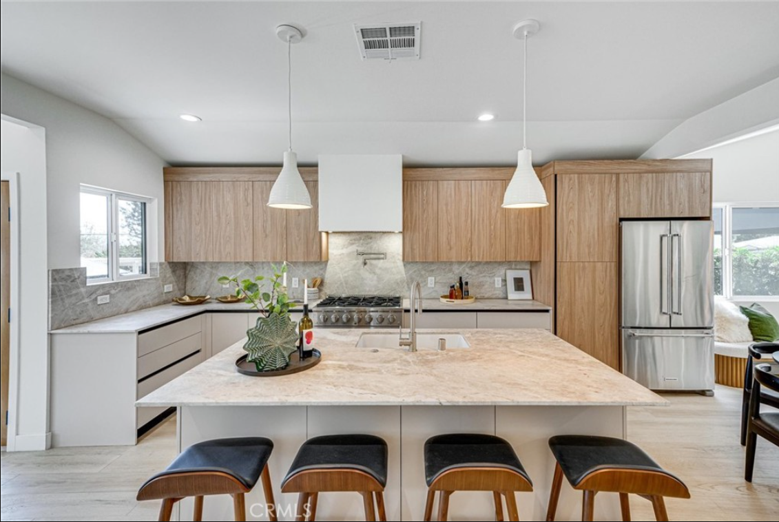 Modern kitchen with light wood cabinets, white range hood, stone island with four stools, and stainless steel appliances.