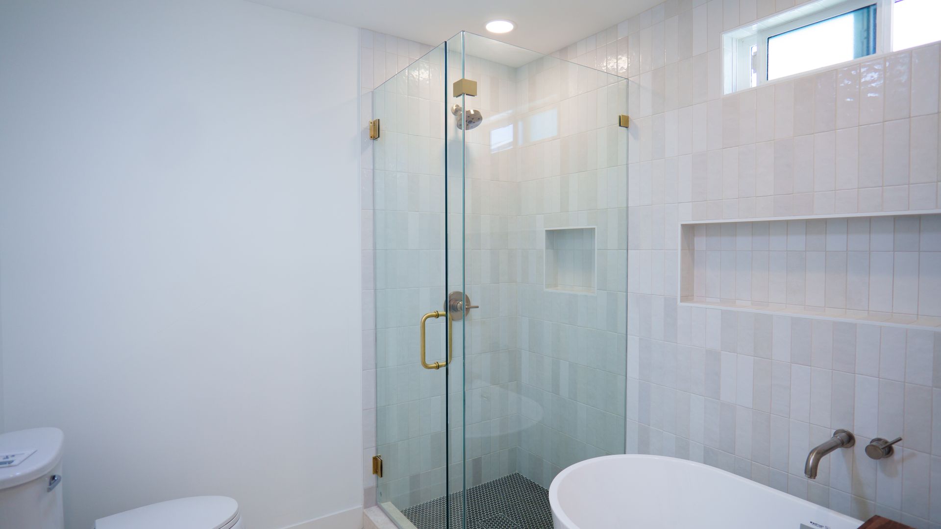 A bright modern bathroom featuring a glass walk-in shower with stone tile, a white bathtub, and a toilet.