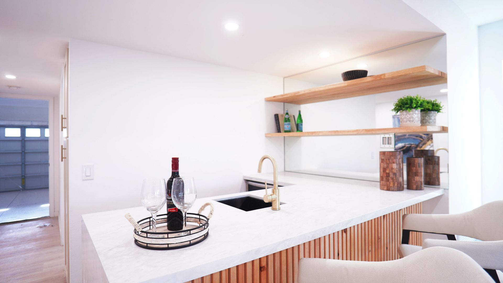A bright kitchen counter with a wine bottle, two glasses, a gold faucet, and wooden cabinets, with shelving on a mirror.