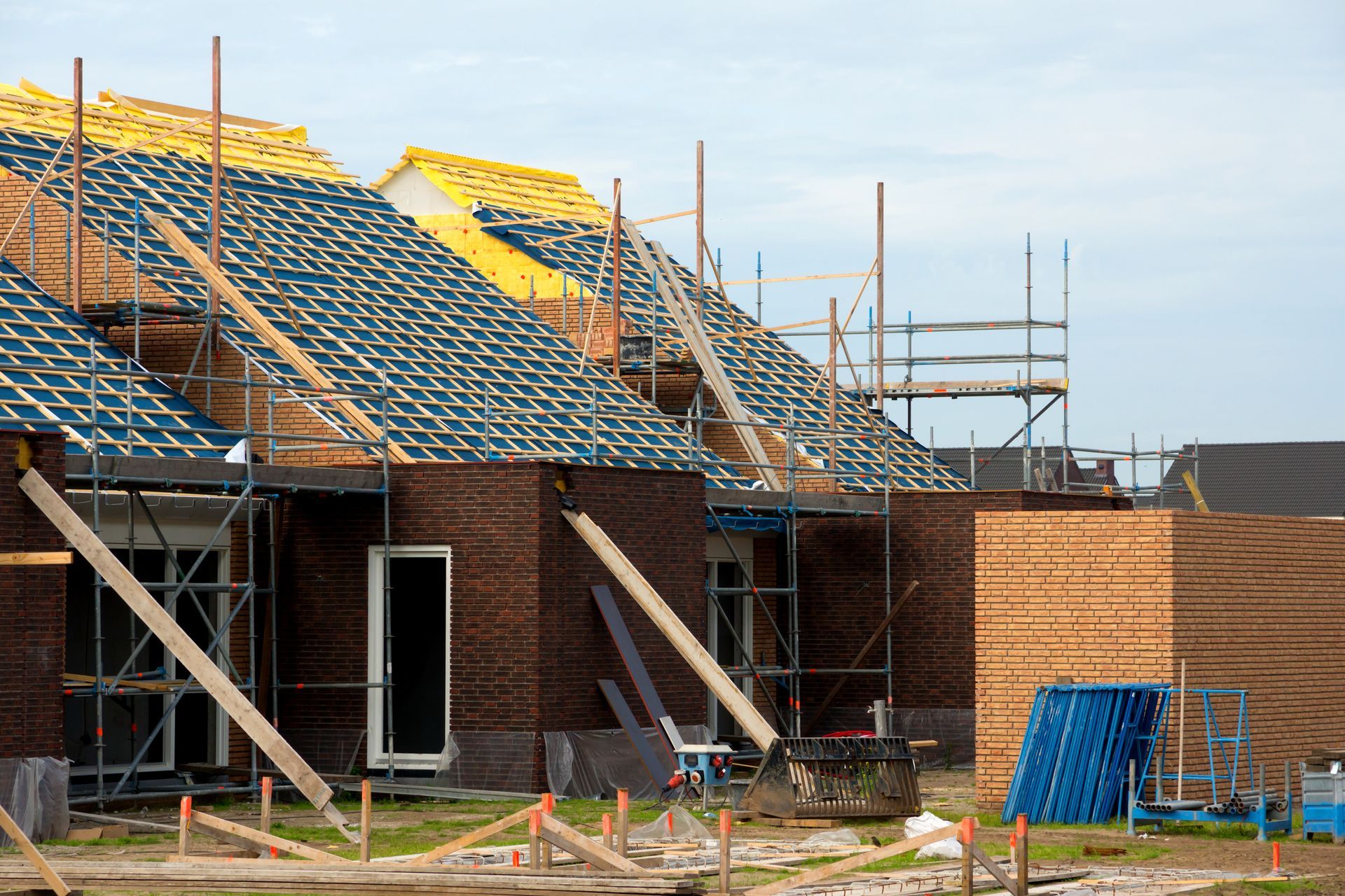 A row of unfinished brick houses under construction with exposed wooden roof frames, scaffolding, and yellow insulation.
