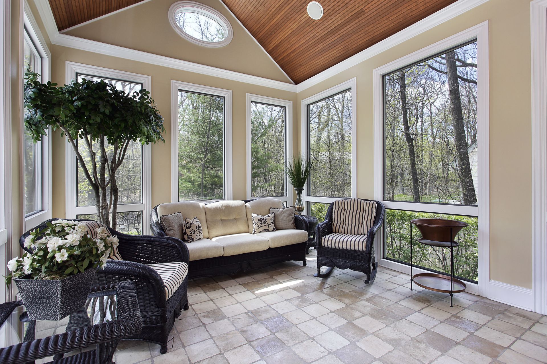 A sunroom with a beige sofa, wicker chairs, a plant, and large windows overlooking trees under a vaulted wood ceiling.