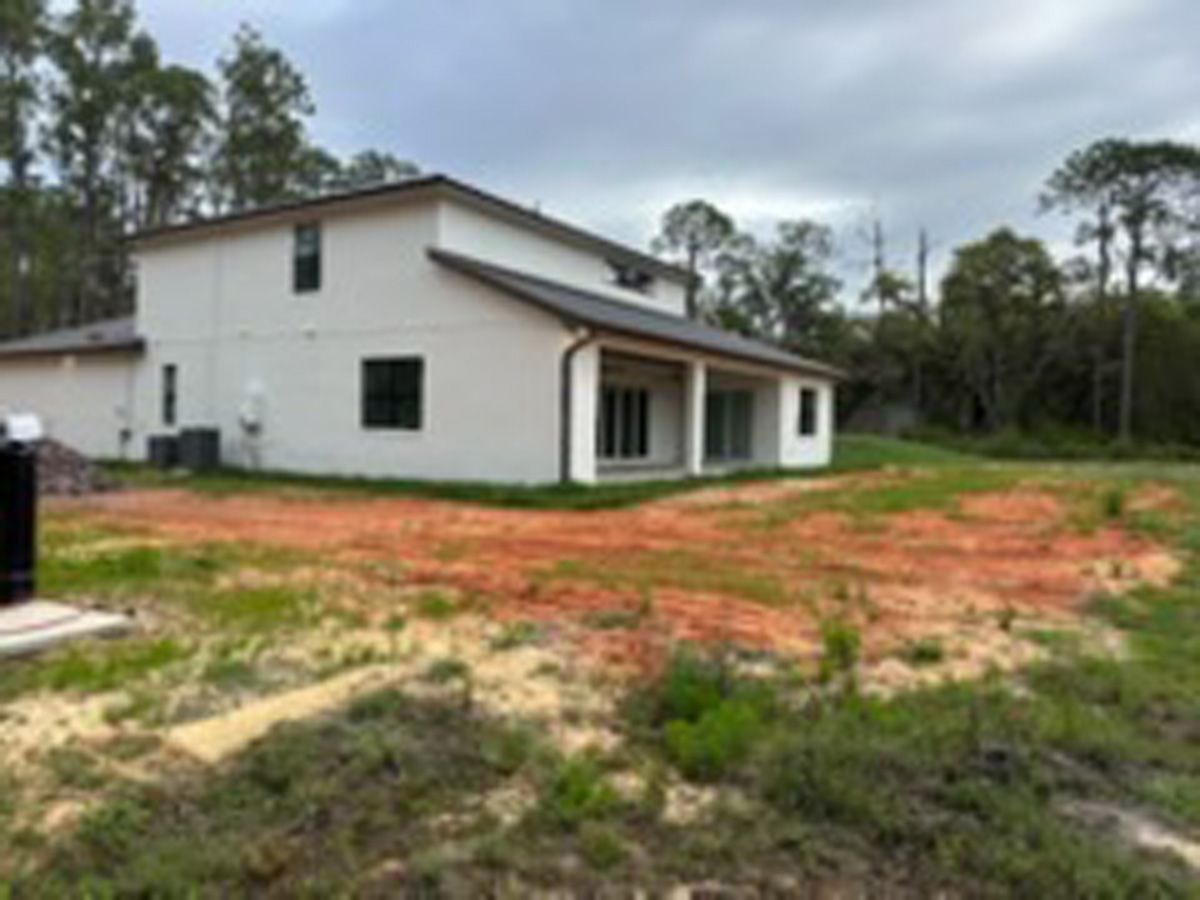 White house with a sloped roof beside a red dirt yard and trees under a cloudy sky